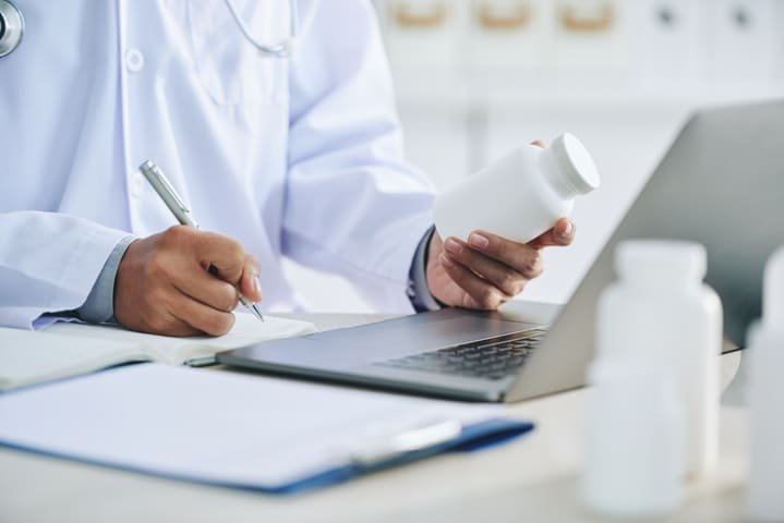 Homem segura frasco de medicamento enquanto anota em um papel com outra mão na frente de um laptop representando registro de medicamentos anvisa